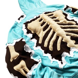 May include: A light blue hoodie with a beige and brown skeleton design on the back. The edges of the design have a white trim. The hoodie has a hood and is made of a soft material.