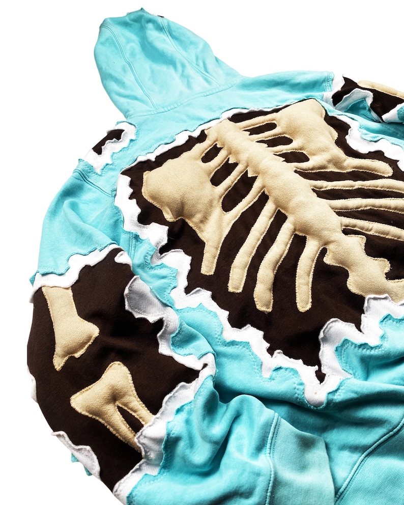 May include: A light blue hoodie with a beige and brown skeleton design on the back. The edges of the design have a white trim. The hoodie has a hood and is made of a soft material.