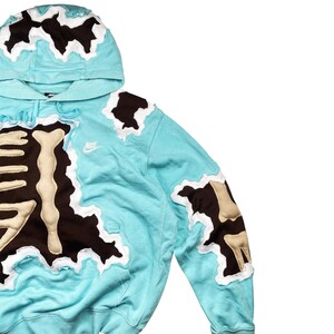 May include: A light blue hoodie with a unique design. The hoodie features brown and beige patches with a white border. The hood and sleeves have similar patches. A small Nike logo is visible on the chest.