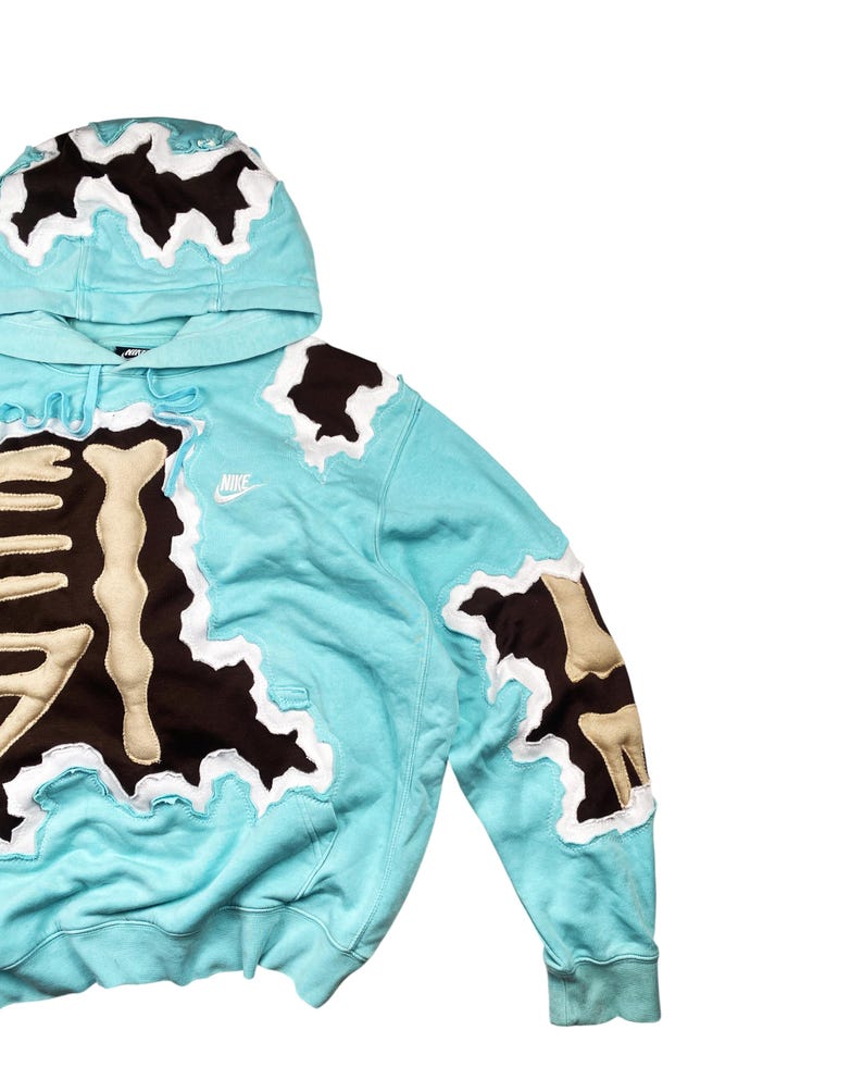 May include: A light blue hoodie with a unique design. The hoodie features brown and beige patches with a white border. The hood and sleeves have similar patches. A small Nike logo is visible on the chest.