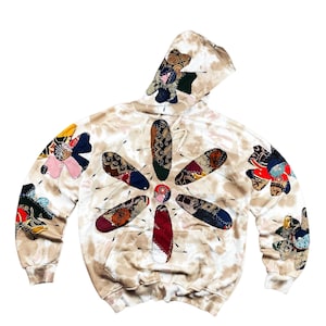 Op de afbeelding: Een beige tie-dye hoodie met een groot bloemenontwerp gemaakt van kleurrijke stoffen patches. De hoodie heeft een capuchon met trekkoord, een voorzak en patches op de mouwen en capuchon.