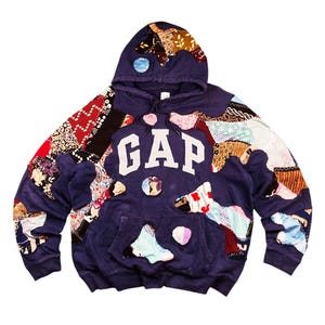 Puede incluir: Una sudadera con capucha GAP azul marino con detalles de patchwork. La sudadera tiene una capucha con cordón, un bolsillo de canguro y el logotipo de GAP en letras blancas. El patchwork incluye varias telas estampadas en rojo, marrón y azul.