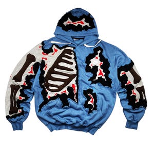 Op de afbeelding: Een blauwe hoodie met een uniek skeletontwerp. Het ontwerp heeft een grijze ribbenkast, bruine botten en rode accenten. De hoodie heeft een capuchon met trekkoord en geribde manchetten.