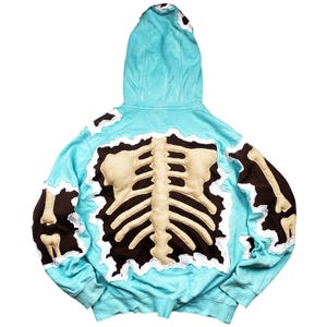 May include: A light blue hoodie with a brown and beige skeleton design on the back. The hoodie has a hood and long sleeves. The skeleton design is made of a beige material and is outlined in brown.