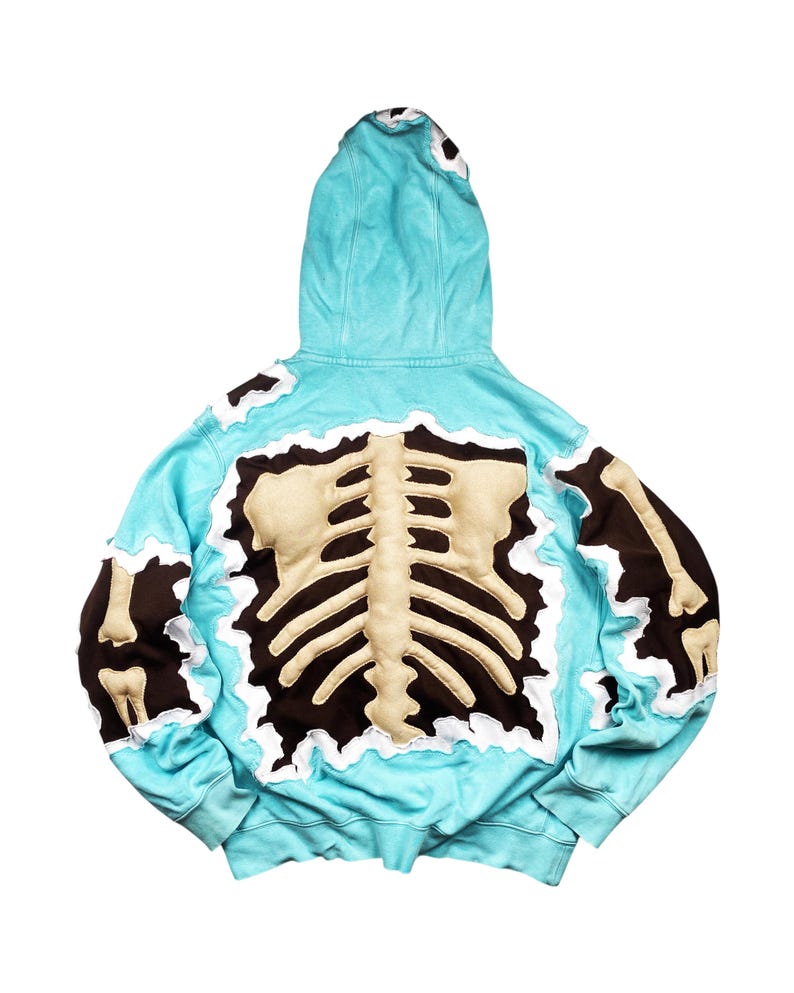 May include: A light blue hoodie with a brown and beige skeleton design on the back. The hoodie has a hood and long sleeves. The skeleton design is made of a beige material and is outlined in brown.