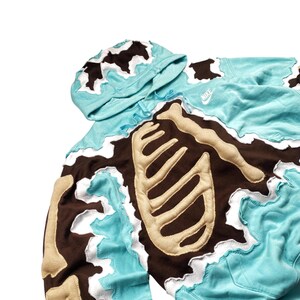 May include: A light blue hoodie with a brown and beige skeleton design. The hoodie features a hood and the Nike logo. The edges of the design have a white, wavy trim. The skeleton design is on the front and sleeves.
