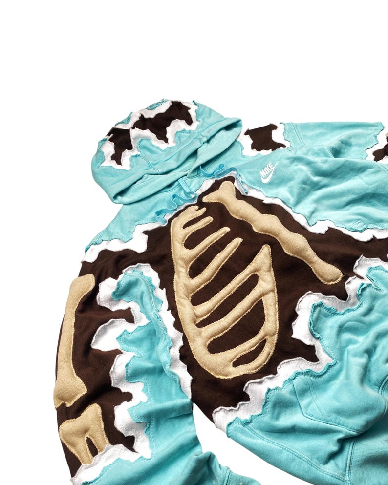 May include: A light blue hoodie with a brown and beige skeleton design. The hoodie features a hood and the Nike logo. The edges of the design have a white, wavy trim. The skeleton design is on the front and sleeves.