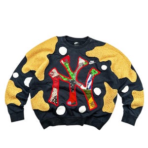 May include: Black Nike sweatshirt with a patchwork design featuring the letters "NY" in red, green, and patterned fabrics. The sweatshirt has yellow, textured accents and white polka dots. A small Nike logo is visible.