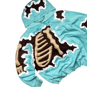 May include: A turquoise hoodie with a skeleton design. The hoodie features a brown and beige skeleton applique over the chest and arms, with white trim. The Nike logo is visible on the chest.