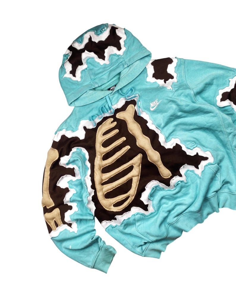 May include: A turquoise hoodie with a skeleton design. The hoodie features a brown and beige skeleton applique over the chest and arms, with white trim. The Nike logo is visible on the chest.