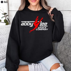 May include: A black long-sleeve crewneck sweatshirt featuring a white and red graphic print on the chest. The design displays the text "abby lee" and "DANCE COMPANY" in white, with a red silhouette of a dancer in a leap pose.