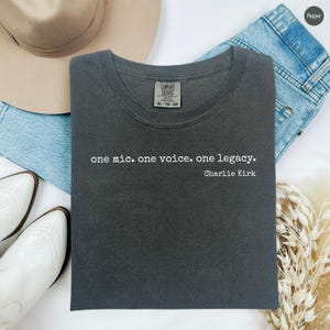 May include: A charcoal gray crew neck t-shirt featuring the white printed text "one mic. one voice. one legacy." followed by "Charlie Kirk" on the front. The shirt has a Comfort Colors brand label on the inside of the collar.