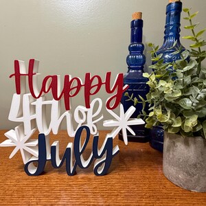 以下が含まれることがあります： 赤、白、ネイビーブルーで「Happy 4th of July」と書かれた木製のサイン。サインはスターバーストで装飾され、木製の表面に置かれています。2本の青いボトルと鉢植えの植物が背景にあります。