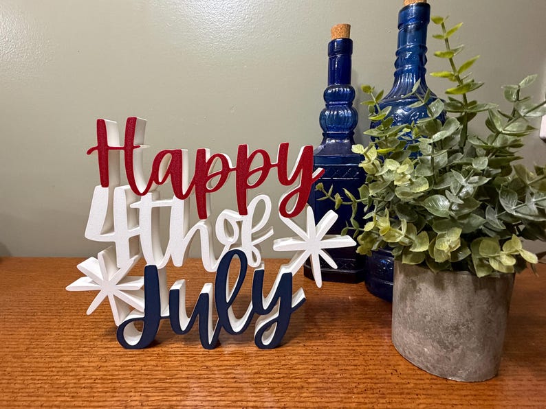 以下が含まれることがあります： 赤、白、ネイビーブルーで「Happy 4th of July」と書かれた木製のサイン。サインはスターバーストで装飾され、木製の表面に置かれています。2本の青いボトルと鉢植えの植物が背景にあります。
