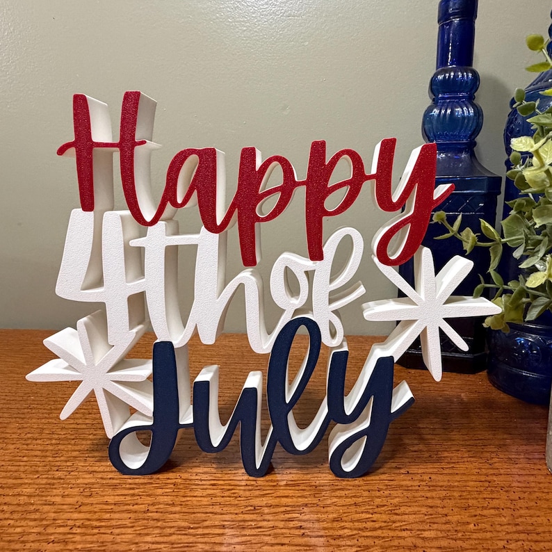 以下が含まれることがあります： 赤、白、紺色の「Happy 4th of July」の文字が書かれた木製のサイン。星型のアクセントがあり、独立記念日の祝賀用にデザインされています。木製の表面に置かれています。