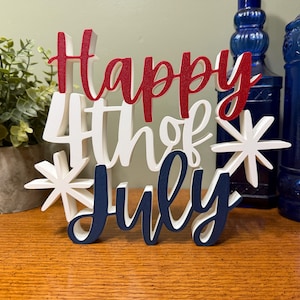 以下が含まれることがあります： 赤、白、青の文字で「Happy 4th of July」と書かれた木製の看板。看板はスターバーストで装飾され、木製の表面に置かれています。背景には青いガラス瓶と鉢植えの植物があります。