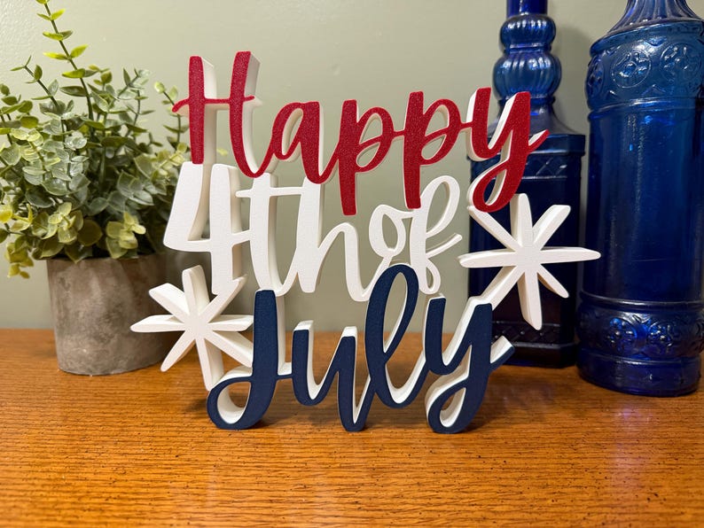 以下が含まれることがあります： 赤、白、青の文字で「Happy 4th of July」と書かれた木製の看板。看板はスターバーストで装飾され、木製の表面に置かれています。背景には青いガラス瓶と鉢植えの植物があります。