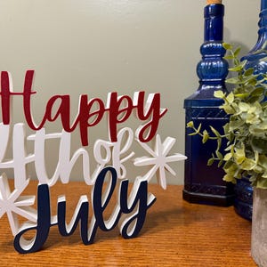 以下が含まれることがあります： 「Happy 4th of July」の文字が赤、白、青で書かれた木製のサイン。サインは星の飾りで飾られ、木製の表面に置かれています。2本の青いガラス瓶と鉢植えの植物が背景に見えます。