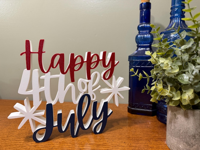 以下が含まれることがあります： 「Happy 4th of July」の文字が赤、白、青で書かれた木製のサイン。サインは星の飾りで飾られ、木製の表面に置かれています。2本の青いガラス瓶と鉢植えの植物が背景に見えます。