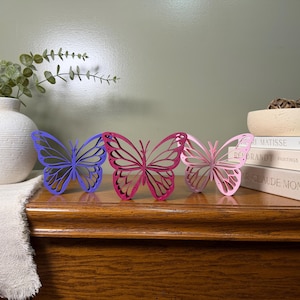 May include: Three freestanding butterfly sculptures arranged in a row. The left butterfly is purple, the center butterfly is magenta, and the right butterfly is light pink. Each butterfly features a layered, intricate cutout wing design.