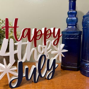 以下が含まれることがあります： 「Happy 4th of July」の文字が赤、白、紺色で書かれた木製のサイン。星のアクセントがあり、2本の青いガラス瓶と鉢植えの植物の前に置かれています。お祝いのデコレーションです。