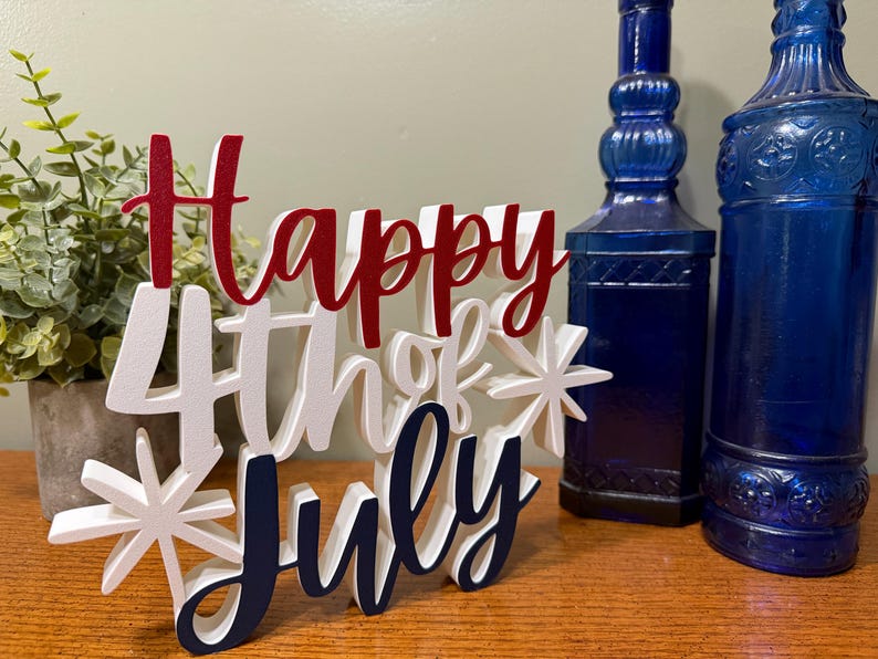 以下が含まれることがあります： 「Happy 4th of July」の文字が赤、白、紺色で書かれた木製のサイン。星のアクセントがあり、2本の青いガラス瓶と鉢植えの植物の前に置かれています。お祝いのデコレーションです。