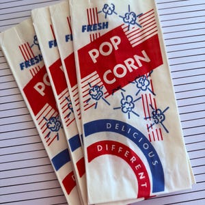 Könnte beinhalten: Vier Vintage-Popcorn-Tüten mit einem rot-weiß-blauen Design. Auf den Tüten stehen die Wörter "FRESH" und "POP CORN". Die Tüten sind auf einer weiß-grau gestreiften Oberfläche angeordnet.