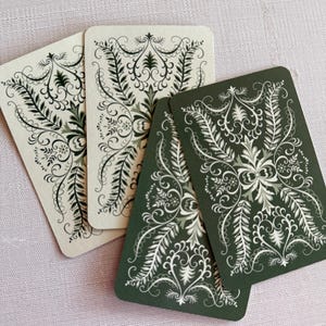 May include: Four rectangular playing cards arranged in a fan shape. Two cards feature a cream background with dark green intricate filigree patterns, while the other two cards display a dark green background with white ornate floral scrollwork.