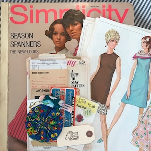 May include: A vintage Simplicity sewing pattern magazine with a blue and white floral fabric flower, a brown and white cat sticker, and a pink and white sewing needle. The magazine cover features the text "Simplicity" and "Season Spanners The New Looks".