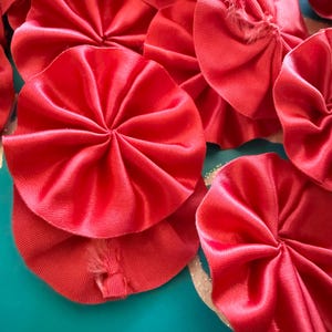 Könnte beinhalten: Nahaufnahme von mehreren leuchtend roten Satin-Stoffrosetten. Die Rosetten sind kreisförmig mit einem plissierten Design, und einige haben ausgefranste Kanten. Der Hintergrund ist türkis.