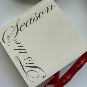 Könnte beinhalten: Ein cremefarbenes Buch mit den Worten "Tis the Season" in eleganter Schrift. Ein rotes Band mit goldenem Text ist unten drapiert. Das Buch ist wahrscheinlich ein Geschenk zum Thema Feiertage.