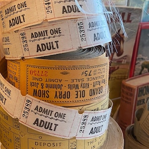 Puede incluir: Una pila de rollos de boletos de papel vintage. Los boletos son de color crema y amarillo con texto negro que dice "ADMIT ONE ADULT". Algunos rollos tienen el texto "GRAND OLE OPRY SHOW".