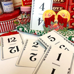 Puede incluir: Tarjetas de números de estilo vintage con números negros y bordes decorativos. También se ve una tarjeta con temática navideña con dos velas encendidas y acentos de acebo. Otros artículos incluyen botes de purpurina, latas a rayas rojas y blancas y chicles.