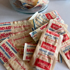 Peut inclure: Une collection de pansements adhésifs individuels de la marque Band-Aid dans des emballages en papier beige. Chaque emballage présente du texte rouge et bleu, incluant le logo Band-Aid, la marque Johnson & Johnson et le nom Sheer Spot.