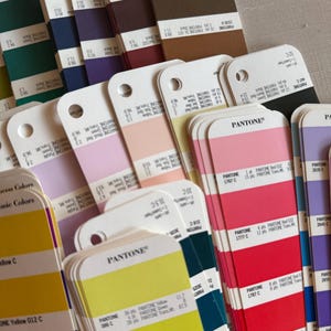 May include: A collection of Pantone colour guide fan decks featuring various swatches in shades of yellow, orange, red, pink, purple, blue, green, brown, and black. Each card displays specific colour names, numerical codes, and ink mixing formulas.