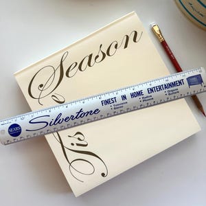 Puede incluir: Un libro crema con la palabra "Season" en escritura elegante. Una regla Silvertone vintage, marcada en centímetros, está sobre el libro. La regla presenta el texto "Finest in Home Entertainment". Un lápiz rojo descansa cerca.
