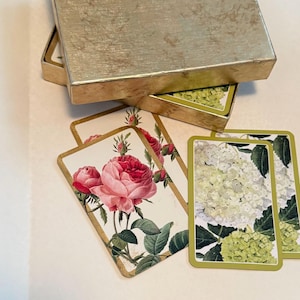 May include: A set of rectangular playing cards with floral illustrations, including pink roses and green hydrangea clusters, featuring gold-toned borders. The cards are partially stored in a matching metallic gold-toned rectangular box with a lid.