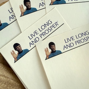 May include: White notecards featuring an image of a man with a raised hand and the text "LIVE LONG AND PROSPER" in black. The man is wearing a blue shirt. The notecards are stacked.