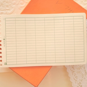 May include: A vintage ledger sheet with a grid pattern and pre-punched holes along the left edge. The sheet has a cream-colored background with light green grid lines. The ledger is resting on an orange envelope.