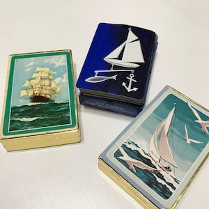 Op de afbeelding: Drie vintage speelkaarten met nautische thema's. Eén kaartspel heeft een zeilschip op een groene achtergrond, een ander heeft een zeilboot en anker op een blauwe achtergrond, en de derde toont een zeilboot met zeemeeuwen op een lichtblauwe achtergrond.