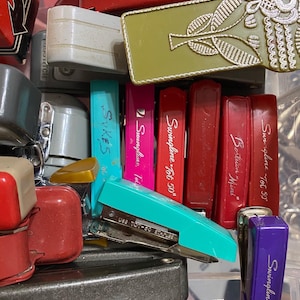 May include: A collection of vintage staplers in various colors, including teal, pink, and red. Some staplers have the brand name "Swingline" printed on them. The image also shows a green and gold decorative piece.