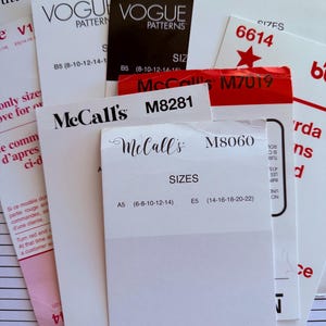 May include: A close-up of various sewing pattern envelopes with different brand names and numbers, including McCall's, Vogue, Simplicity, and Butterick. The envelopes are white with black and red text.