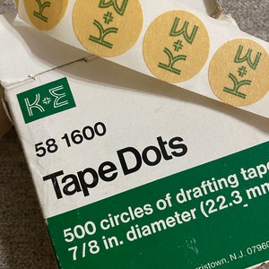 Op de afbeelding: Een rol met 500 zelfklevende tape-stippen, met een diameter van 22,3 mm, in een witte doos met groene en zwarte tekst. De doos heeft de tekst "K+E 58 1600 Tape Dots 500 cirkels van tekentape 7/8 inch diameter (22,3 mm) Keuffel & Esser Company, Morristown, N.J. 07960".