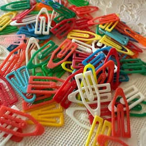 May include: A collection of colorful plastic hair clips in various shapes and sizes. The clips are in shades of red, yellow, green, blue, and white. The image shows a close-up view of the clips scattered on a textured surface.
