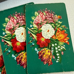 May include: Two rectangular playing cards with a dark green background featuring a central floral arrangement. The bouquet includes pink roses, white blossoms, red flowers, yellow and red striped tulips, and small white lily of the valley sprigs.