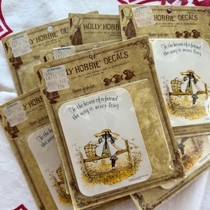 Puede incluir: Una colección de calcomanías de Holly Hobbie en su embalaje original. Las calcomanías presentan a una niña con gorro y vestido, con el texto "To the house of a friend the way is never long". El embalaje es de color canela con etiquetas de precio de JCPenney.