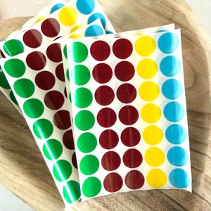Op de afbeelding: Vellen kleurrijke stipstickers in groen, bordeaux, geel en blauw. De stickers zijn in rijen gerangschikt op een witte achtergrond. De stickers liggen in een houten kom.