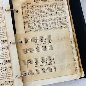 May include: A vintage hymnal with a black binder and pages filled with sheet music. The music is titled "Jesus is Calling Thee" and includes lyrics and musical notation. The hymnal is open to a page with the words "Now can I help" written in cursive.