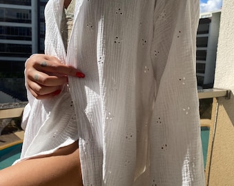 May include: White, open-front cover-up with floral eyelet details. The lightweight fabric has a textured appearance. The cover-up is worn over a green top. The background includes a building and a sunny sky.