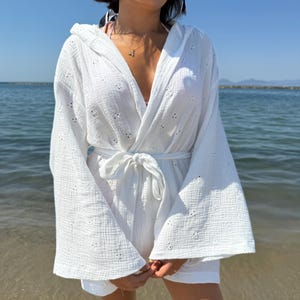Könnte beinhalten: Weißer Strand-Cover-up mit Kapuze und Bindegürtel. Der Cover-up hat ein strukturiertes, perforiertes Design und weite Ärmel. Die Person trägt eine Sonnenbrille und eine Halskette. Der Hintergrund ist ein Strand mit blauem Wasser und klarem Himmel.
